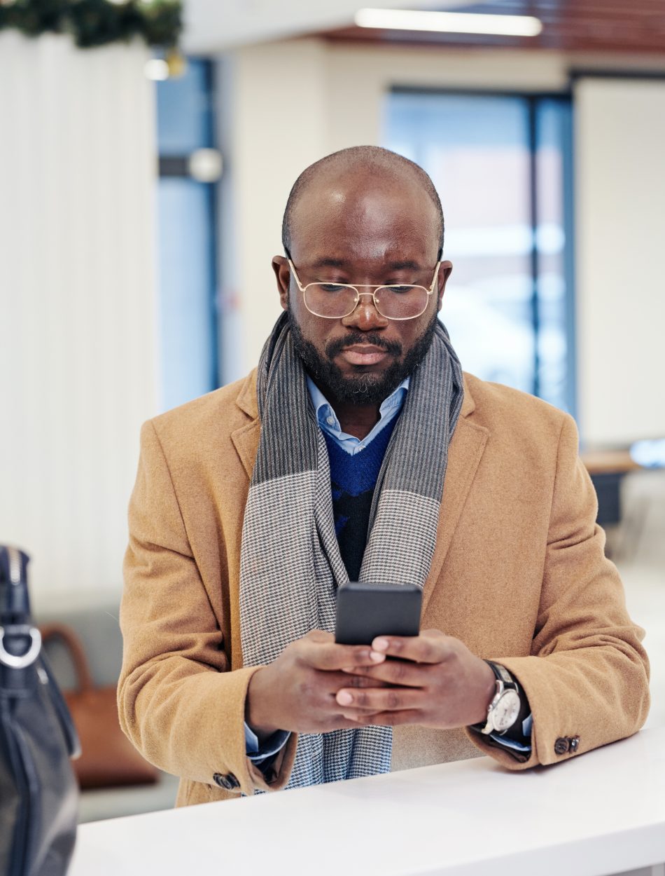 Serious African businessman in coat reading a message on mobile phone while standing near the reception at the lobby